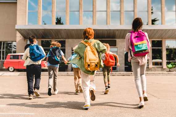 Kinder gehen mit Rucksäcken auf eine Schule zu.