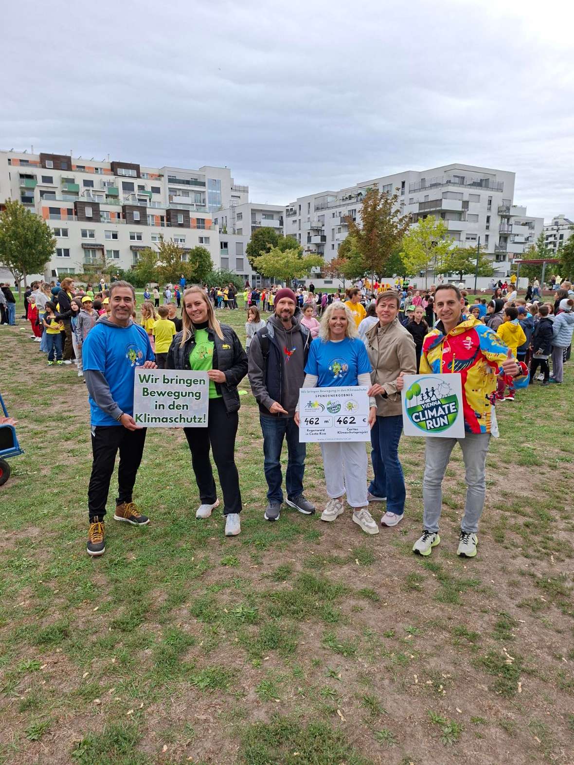 5 Personen halten 3 Schilder in die Höhe auf denen steht: "Climate Run" und "Wir bringen Bewegung in den Klimaschutz". Im Hintergrund sieht man viele Kinder auf einer Wiese.